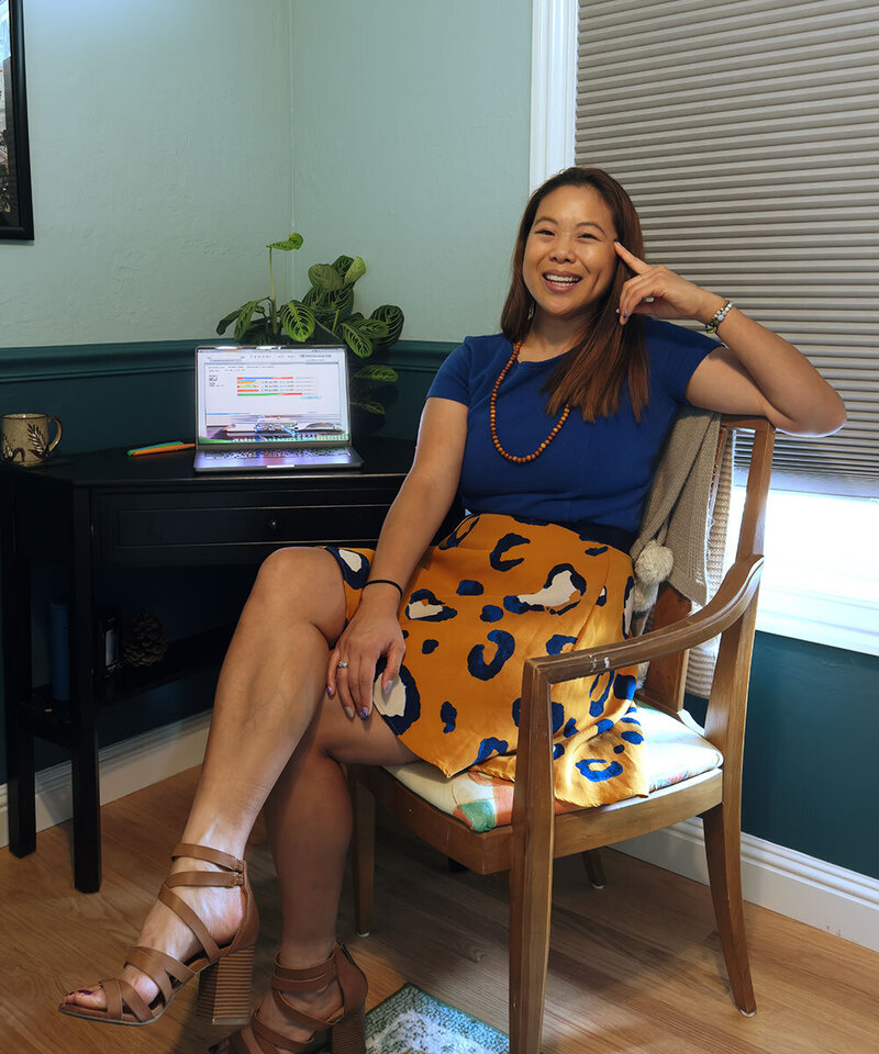 Asian-American woman wearing blue top and orange patterned skirt is sitting in a chair.