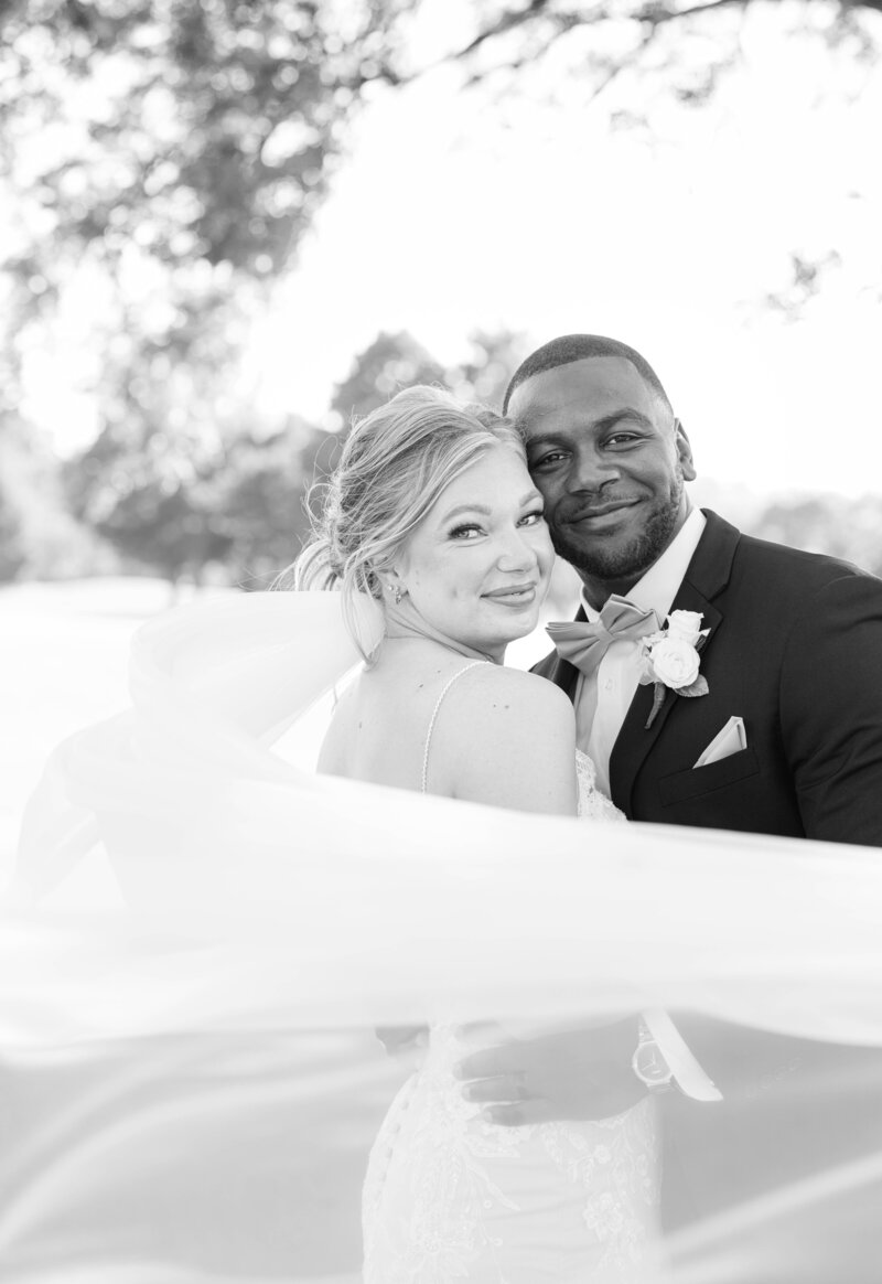 A black and white photo of a bride and groom with her veil  blowing in the wind