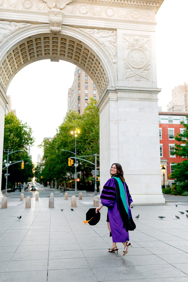 NYU graduate smiling and walking through the Washington Square Park Arch at sunrise