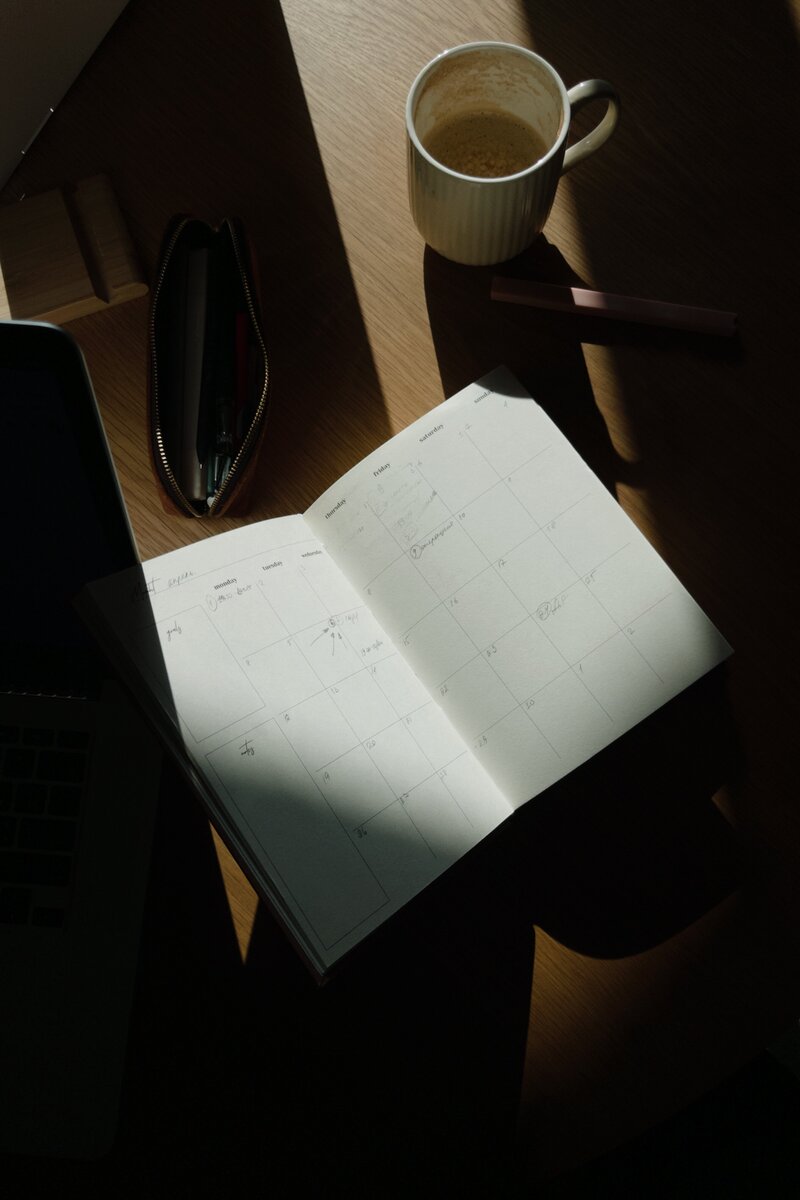 An open planner and a mug of coffee bathed in sunlight sit on a wooden table. Nearby, a pen case and a laptop add to the cozy, productive scene.