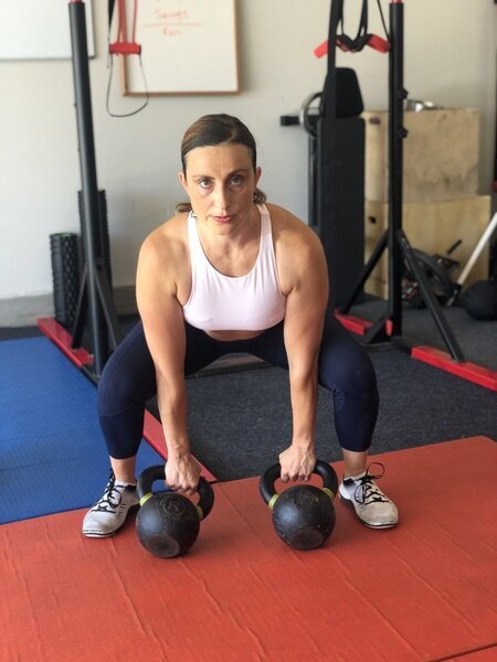 Vanessa Flores in a home gym bending at the hips and knees to grip two kettlebells on an orange mat, surrounded by gym equipment, preparing for a strength training lift