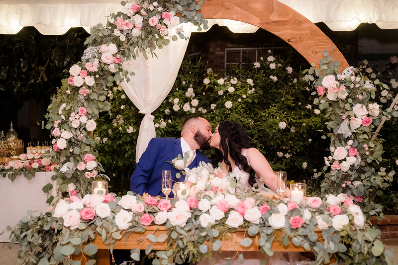bride and groom share a kiss while they set at their wedding table surrounded by wedding florals