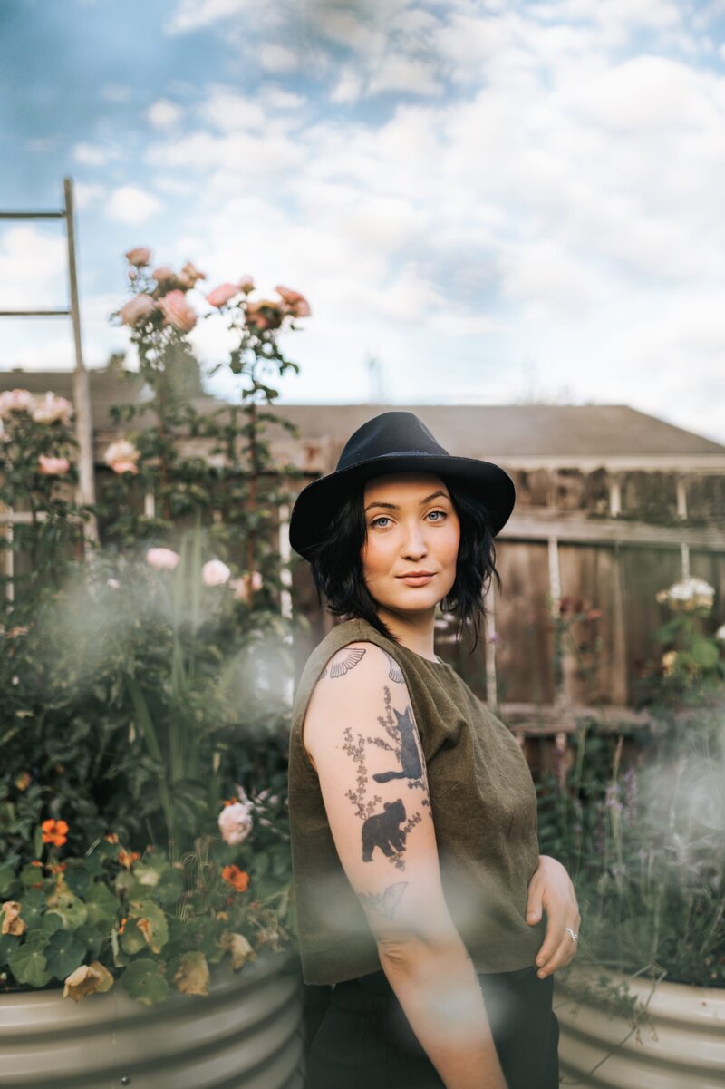 Woman with tattoos wears a black hat and dark green sleeveless top standing outside in a garden and softly smiling at the camera.