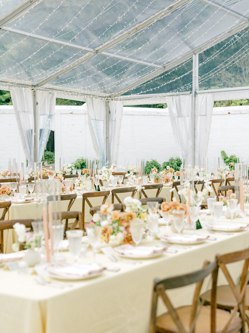 Al fresco wedding reception table in a green clearing with a white textured linen, wooden chairs, and bright colorful flower centerpieces and candles on top