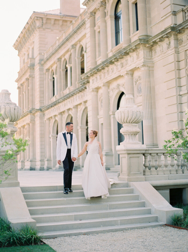 Rhode Island Wedding Photographer | A couple in formal wedding attire, holding hands, walks down stone steps in front of an elegant, historic building.