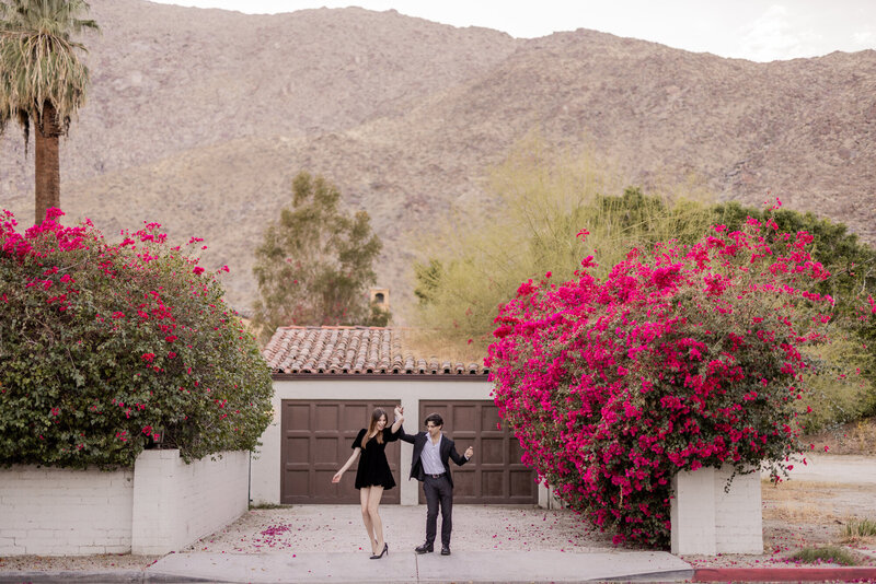 "A fashionable couple dances in a dreamy Palm Springs setting, surrounded by vibrant pink bougainvillea and classic Spanish-style architecture. With the desert mountains as a backdrop, this editorial-style portrait captures a romantic and effortlessly chic moment.