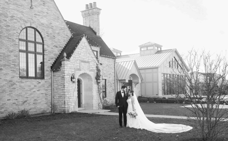 A black and white photo of a bride and groom standing outside a brick manor on their wedding day in the fall.