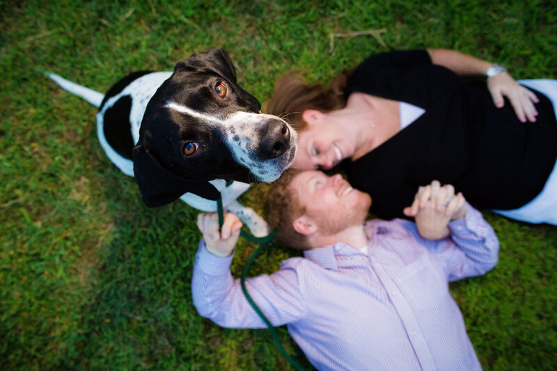 A playful moment captured during an engagement session with a dog looking up at the camera while the engaged couple lies out of focus on the ground, showcasing a creative perspective and the couple's love for their furry family member.