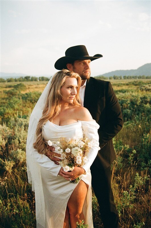 A couple stands together in a field at their wedding in Jackson, Wyoming. One partner, wearing a white off-the-shoulder dress, holds a bouquet of wildflowers, while the other, dressed in a black suit and cowboy hat, stands protectively behind them, gazing into the distance.