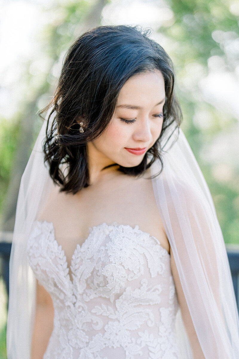 This radiant bride, captured in the soft California sunlight, exudes timeless elegance. Her lace gown features intricate floral detailing, paired perfectly with a delicate veil that cascades gracefully. The serene outdoor backdrop enhances the bride’s natural beauty, making this moment a quintessential part of her California wedding. Keywords: California wedding, lace gown, elegant bride.