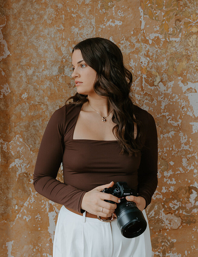 Wedding photographer, Stevie Davis, in a brown top and white pants holding a camera. She gazes to the side, against a textured, rustic wall backdrop.