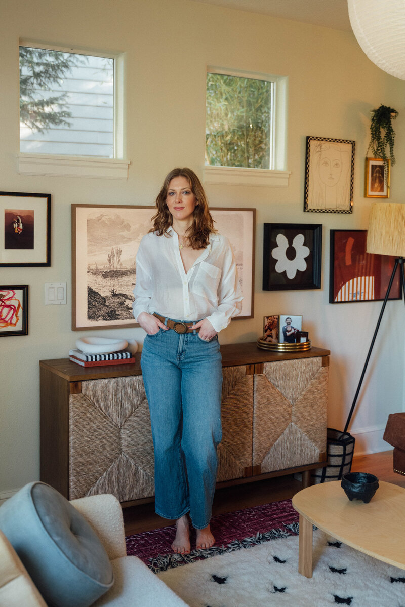 Erin Lima, an interior designer in Portland and Lake Oswego, stands with her hands in her jean pockets in front of a wall featuring artwork in her living room. 
