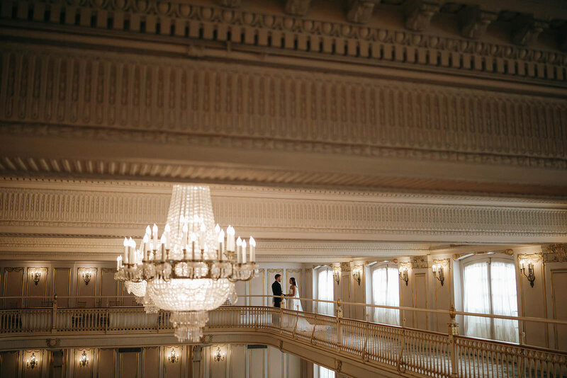 Bride and groom holding hands on a grand ballroom balcony beneath a crystal chandelier, photographed by a Washington wedding photographer.