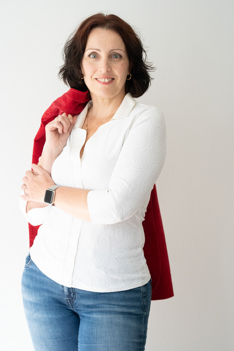 Csilla standing, and holding her red jacket over her shoulder, with a slight smile.