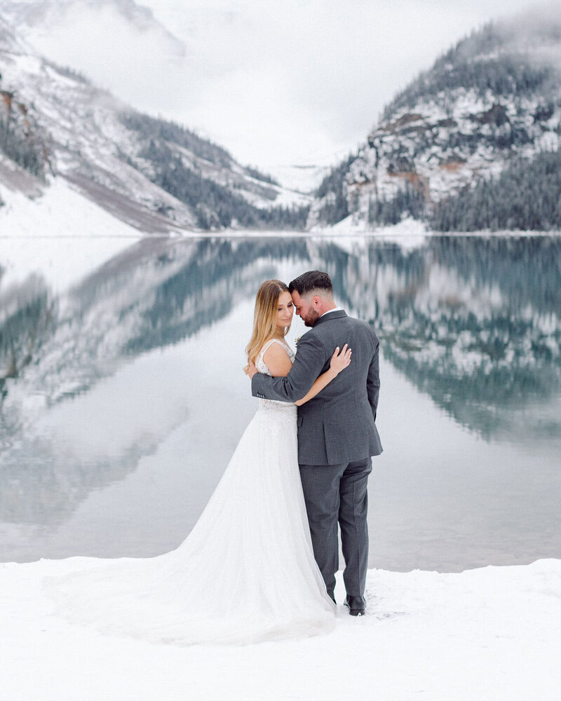 A stunning winter photograph of a bride and groom at Lake Louise after their Deer Lodge wedding, with snow-covered surroundings and the serene, flat lake reflecting the beauty of the day.
