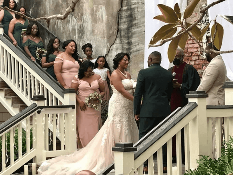 A close up of bride and groom stand facing each other during their wedding ceremony on an outdoor staircase, surrounded by bridesmaids in pink dresses.