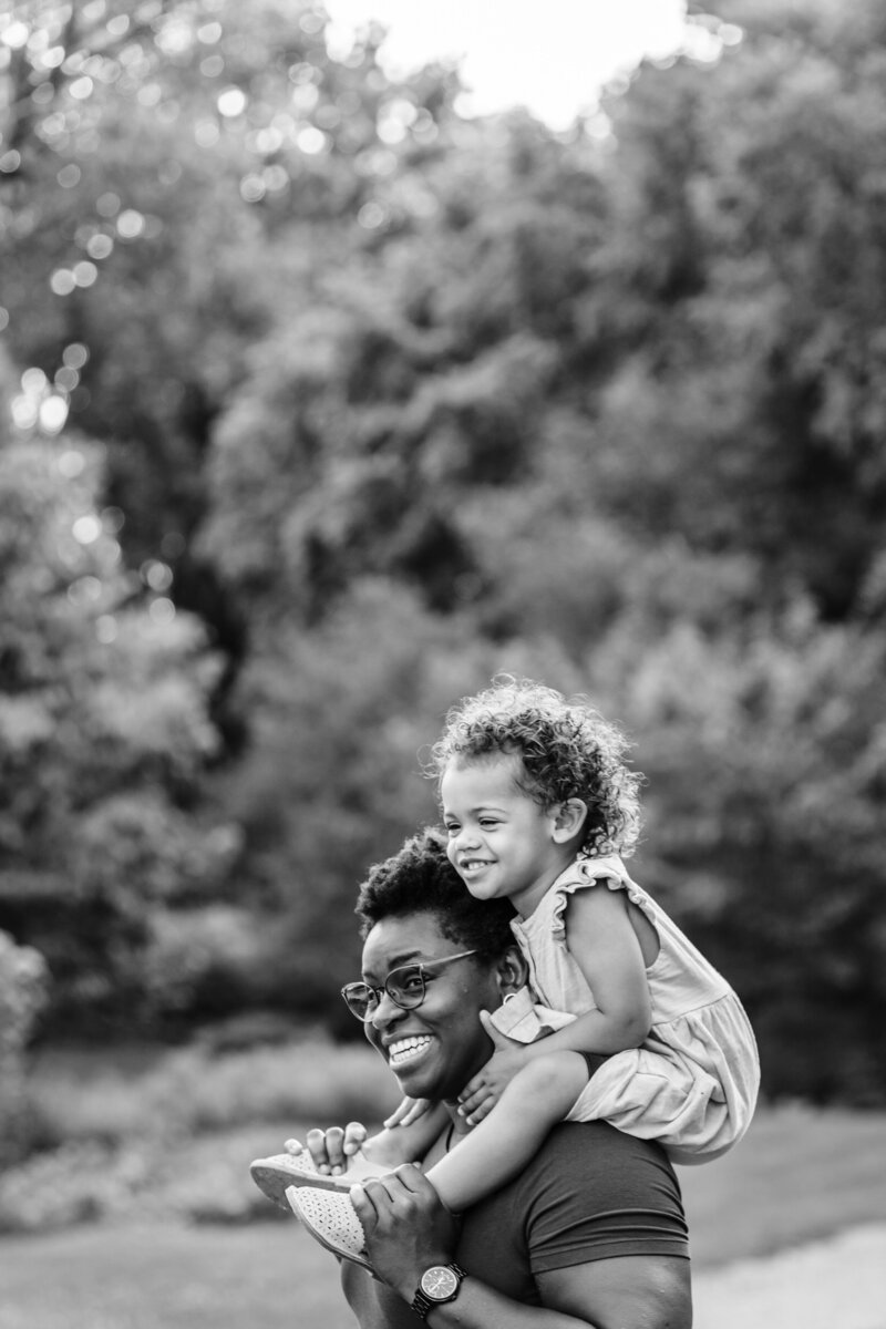 A black and white image of a parent carrying their child on their shoulders while walking in a park. The child is smiling joyfully, and both seem to be enjoying the moment.