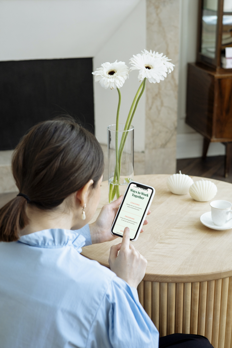 A person seated at a wooden table, browsing coaching programs on a smartphone with a vase of white flowers in the background.