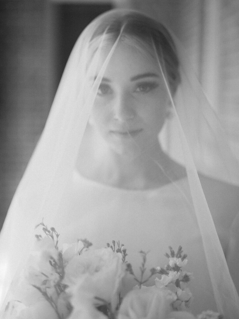 Black and white portrait of a bride under her veil, holding a bouquet of flowers, with a soft, ethereal expression.