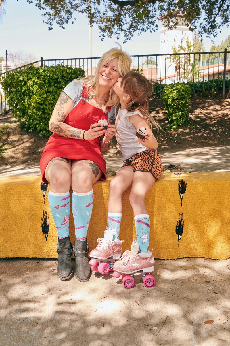 A blonde tattooed woman wearing a red dress leans into her daughter for a kiss. They are wearing matching socks and the daughter has pink rollerskates on.