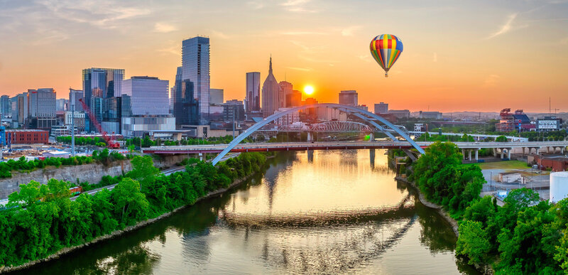 A vibrant cityscape at sunset with a hot air balloon in the sky. The sun is setting behind tall buildings and a distinctive arched bridge over a river, reflecting the warm colors of the sky. Lush greenery lines the riverbanks.