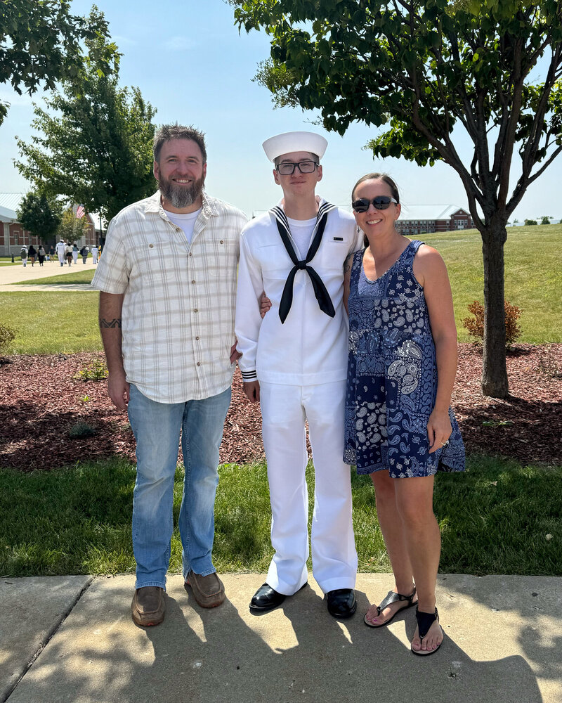 3 people standing next to each other outside on a sunny day.  Father, son, and step-mom.  Father wearing blue jeans and a short sleeve button up creme shirt with brown shoes, beard, and short brown hair.  Son is wearing a US Navy suite that is white with a black tie and black shoes.  Mom wearing a short blue dress, brunette, black shoes and wearing dark sunglasses.