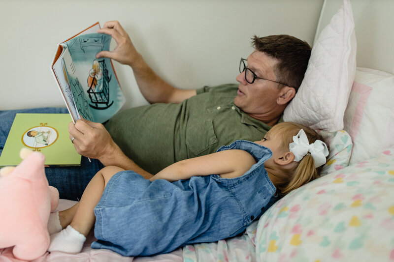 little girl lays next to her dad in her bed while he reads her a book.