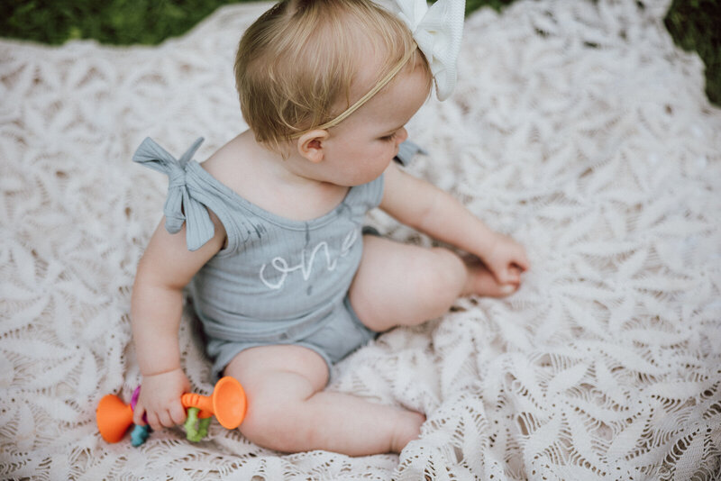 Portrait taken of a one year old girl sitting on a lace blanket holding a toy in one hand and looking at her other hand that is playing with her toes. 