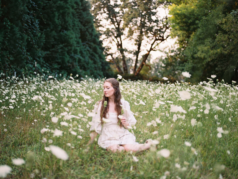 Eugene Wedding Photographer, Heather, sits in a field of white wildflowers, representing her love for documenting nature and mundane moments. 
