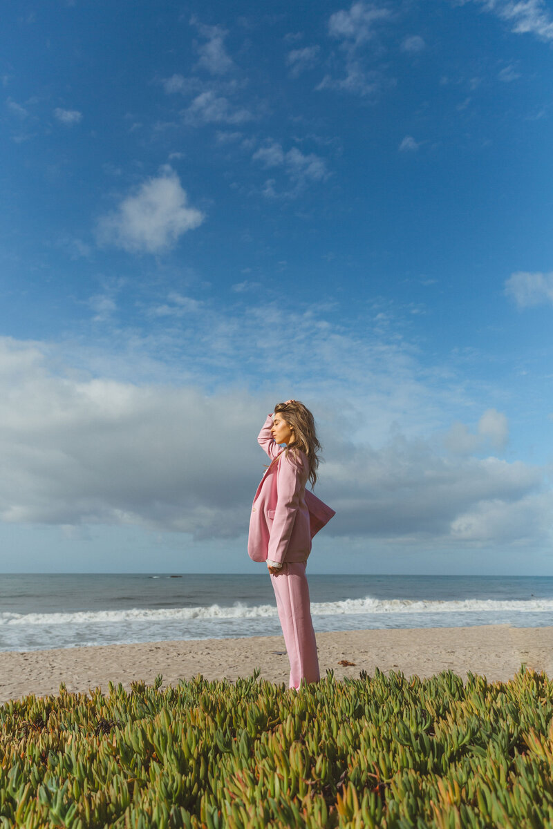 Jessa Jones, the owner of Shindig Social, standing on a beach in San Clemente, California, wearing a pink suit and sunglasses. The ocean and a cloudy sky are in the background. The text overlay reads 'Faith Building,' highlighting Shindig Social's emphasis on faith and community.