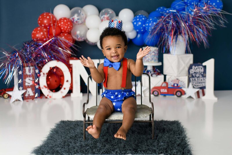 little boy wearing all americana colors sits and smiles in his custom july fourth birthday cake smash session