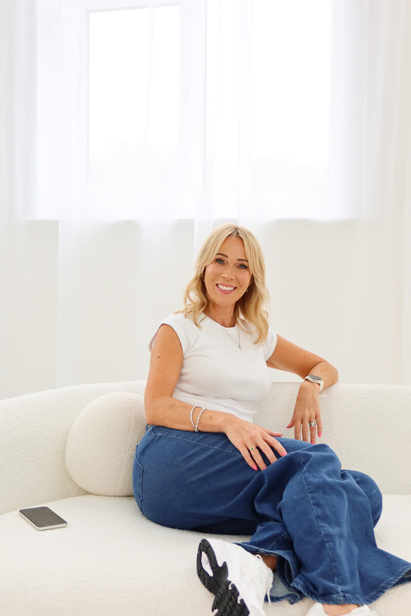 A woman with blonde hair, wearing a white top and blue jeans, sits smiling on a white sofa. A smartphone rests beside her, and the background features white curtains and bright natural light.