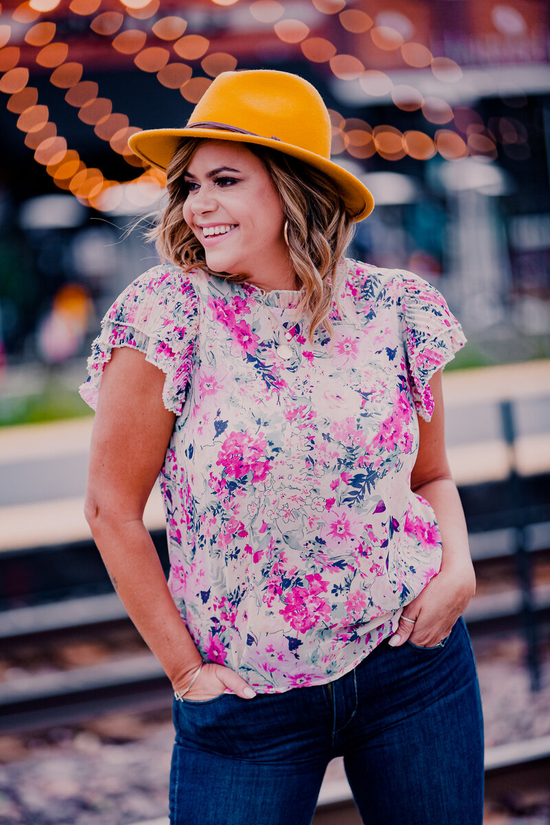 Light skinned woman wearing a yellow fedora hat, floral shirt and jeans standing in front of a blurred outdoor background.