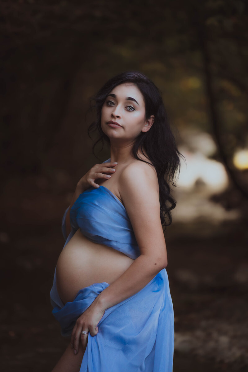 A maternity photo in the woods of a  mother in  blue chiffon wrapped around her body that only shows her face, arms, and baby bump