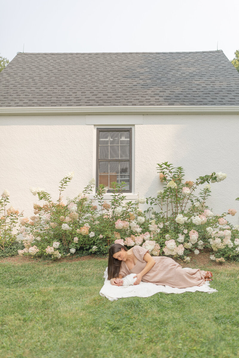 A mother lays on a soft blanket in the grass, gazing lovingly at her swaddled newborn during a serene outdoor newborn session. The natural setting and warm light create a peaceful and intimate moment between mother and baby