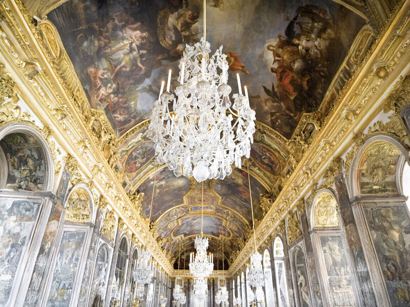 Lavish hall with ornate gold details and large chandeliers hanging from a painted arched ceiling. The walls are lined with tall mirrors and intricate artwork, creating an elegant and grand atmosphere.