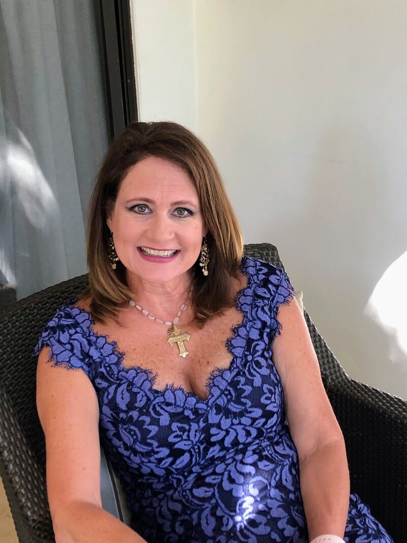 A portrait of a destination wedding planner wearing a lace-patterned blue dress, sitting in a chair and smiling. She is accessorized with jewelry and photographed in an outdoor setting