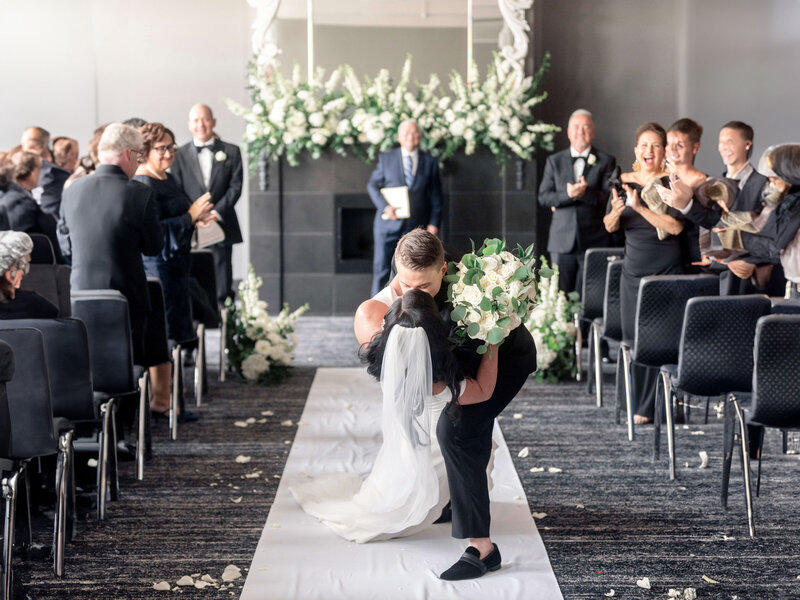 A couple kisses at the end of a wedding aisle, surrounded by cheering guests in formal attire. The venue is elegantly decorated with white flowers and a black backdrop. The bride holds a bouquet of white roses.