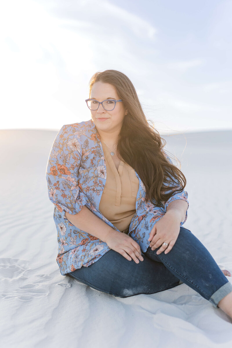A Dayton Wedding Photographer sits on the sand in smiling at the camera. She is wearing a blue shirt with flowers on it and jeans.