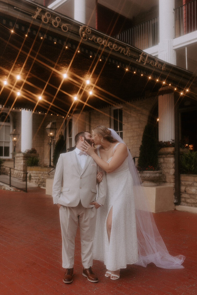 A bride and groom in wedding attire stand in front of the Crescent Hotel in Eureka Springs, Arkansas. They are kissing in front of the awning that says "1886 Crescent Hotel" in ornate gold lettering. The lights underneath the awning create a magical starburst effect.