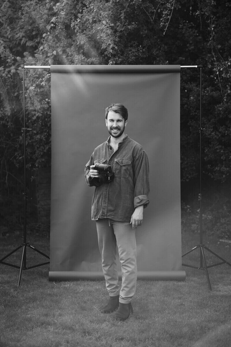 Black and white portrait of Tom Keenan holding a camera, standing in front of a backdrop outdoors.