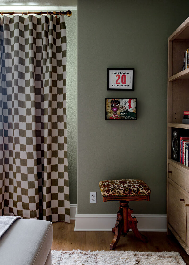  A corner of a room showing checkered curtains, fun art work on the wall and a stool with printed fabric, representing maximalist interior design.
