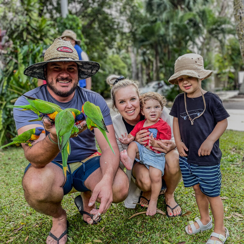 A family on holiday with two little children holding many bright coloured birds on their arms. 