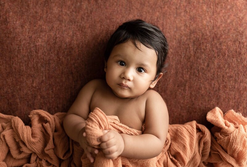 Baby Photoshoot in London, Ontario Studio baby is laying on rust-coloured blanket and draped in coordinating fabric blanket. Aerial image - baby is looking intently at the camera.