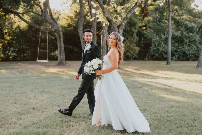 A bride in a strapless white gown and groom in a dark suit walk through a grassy, sun-dappled outdoor setting with trees and a distant swing.