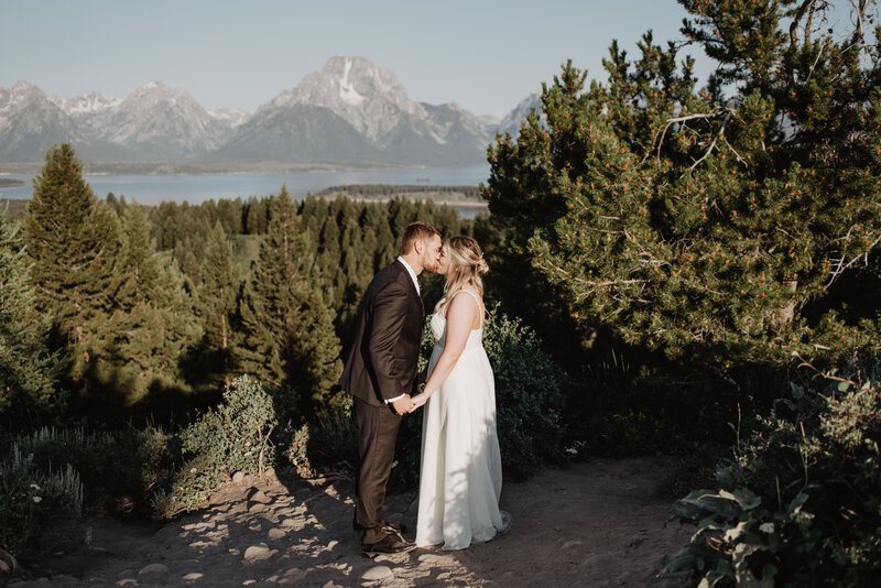 jackson hole wedding with bride and groom holding hands and kissing as they stand on an outlook together with the grand tetons in the distance through the forest