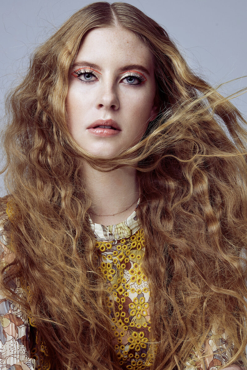 Close-up editorial-style portrait of a teen model with long, voluminous wavy hair wearing a retro yellow crochet top, captured in a studio with soft lighting.
