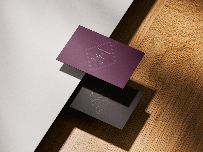 A purple and black business card set with "Transformation with GRIT & GRACE" and a minimalist snake logo, placed on a wooden surface.