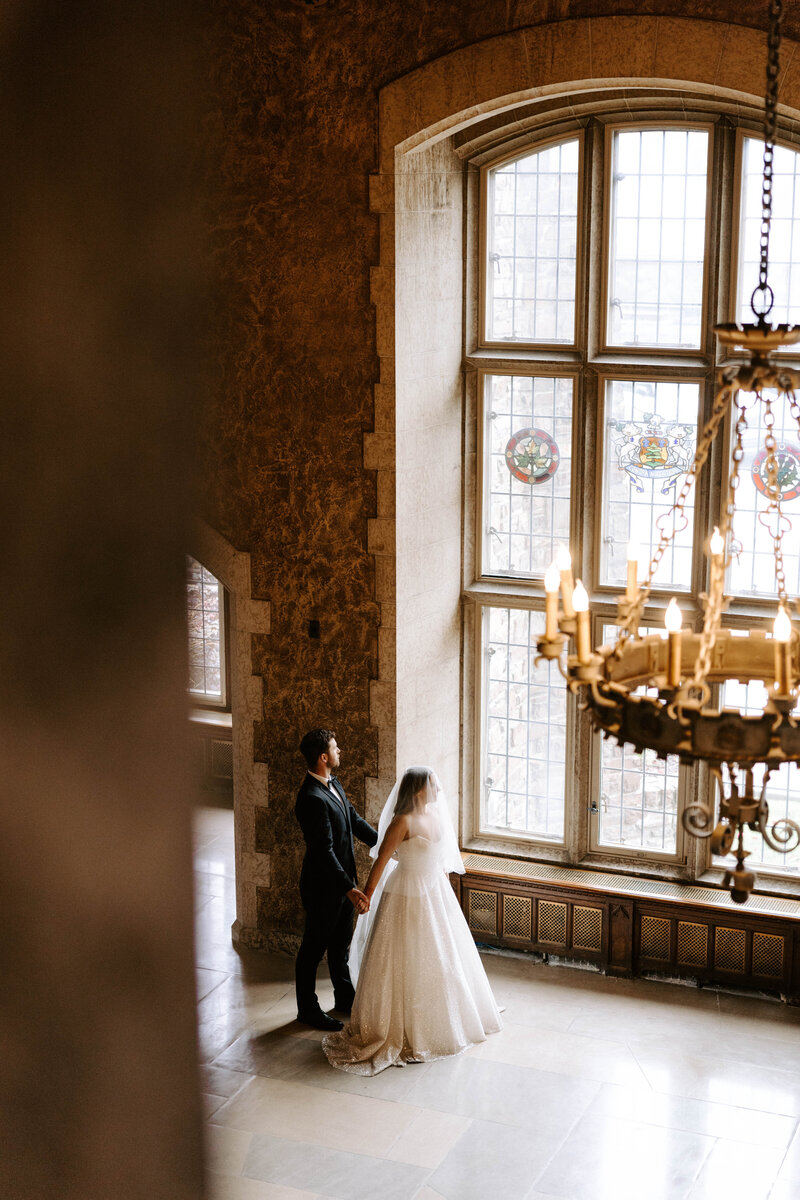 Bride and groom starring out beautiful stain-glass window.  Candlelit chandelier, stonework  Banff, Alberta