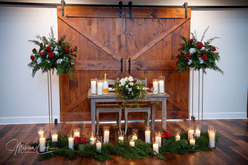 A rustic wedding setup showcases an elegant reception venue gallery with a wooden table adorned with floral arrangements and candles. Two large floral stands flank the table by wooden sliding barn doors, creating charming barn wedding pictures Virginia amidst the greenery and candlelit glass holders.